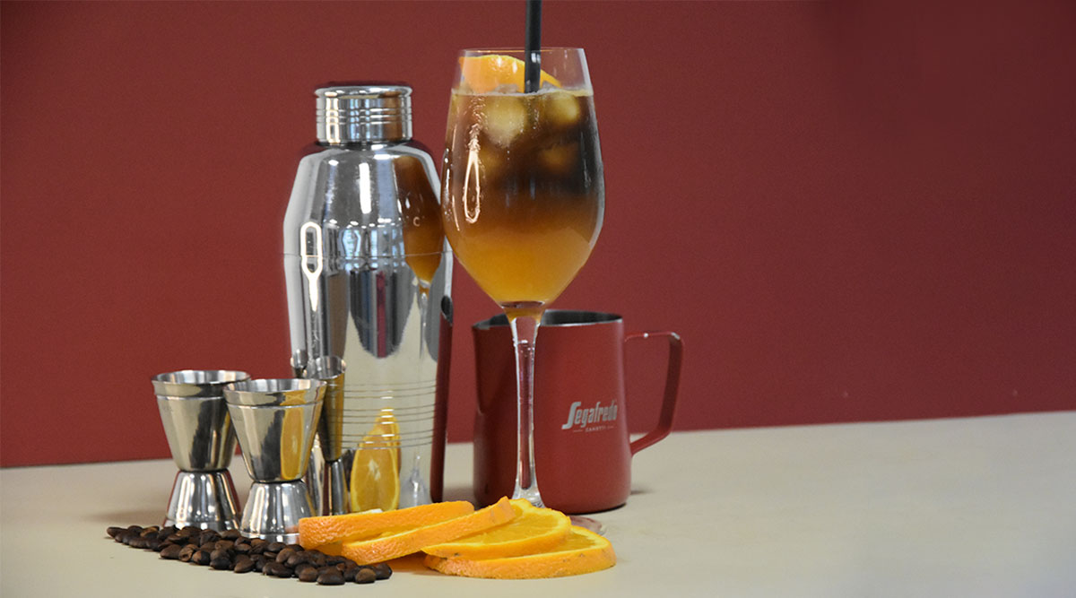 Ein Weinglas gefüllt mit Orangensaft und Kaffee, dekoriert mit einer Orangenscheibe steht neben einem Cocktailshaker.