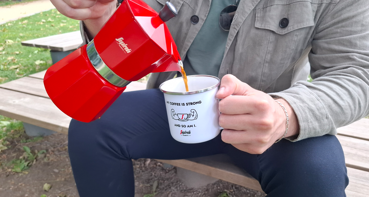 Ein Mann sitzt auf einer Bank im Freien und schenkt sich Kaffee aus einer Mokakanne ein
