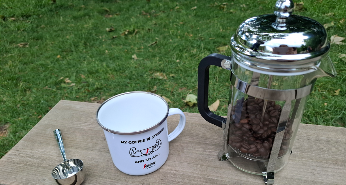 Eine Emaille-Tasse, ein Kaffeemaß und eine French Press stehen auf einem Brett auf einer Wiese