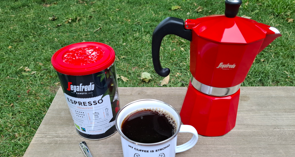 Eine Dose mit Kaffee, ein Emaille-Becher und eine Moka Kanne stehen auf einem Brett auf einer Wiese