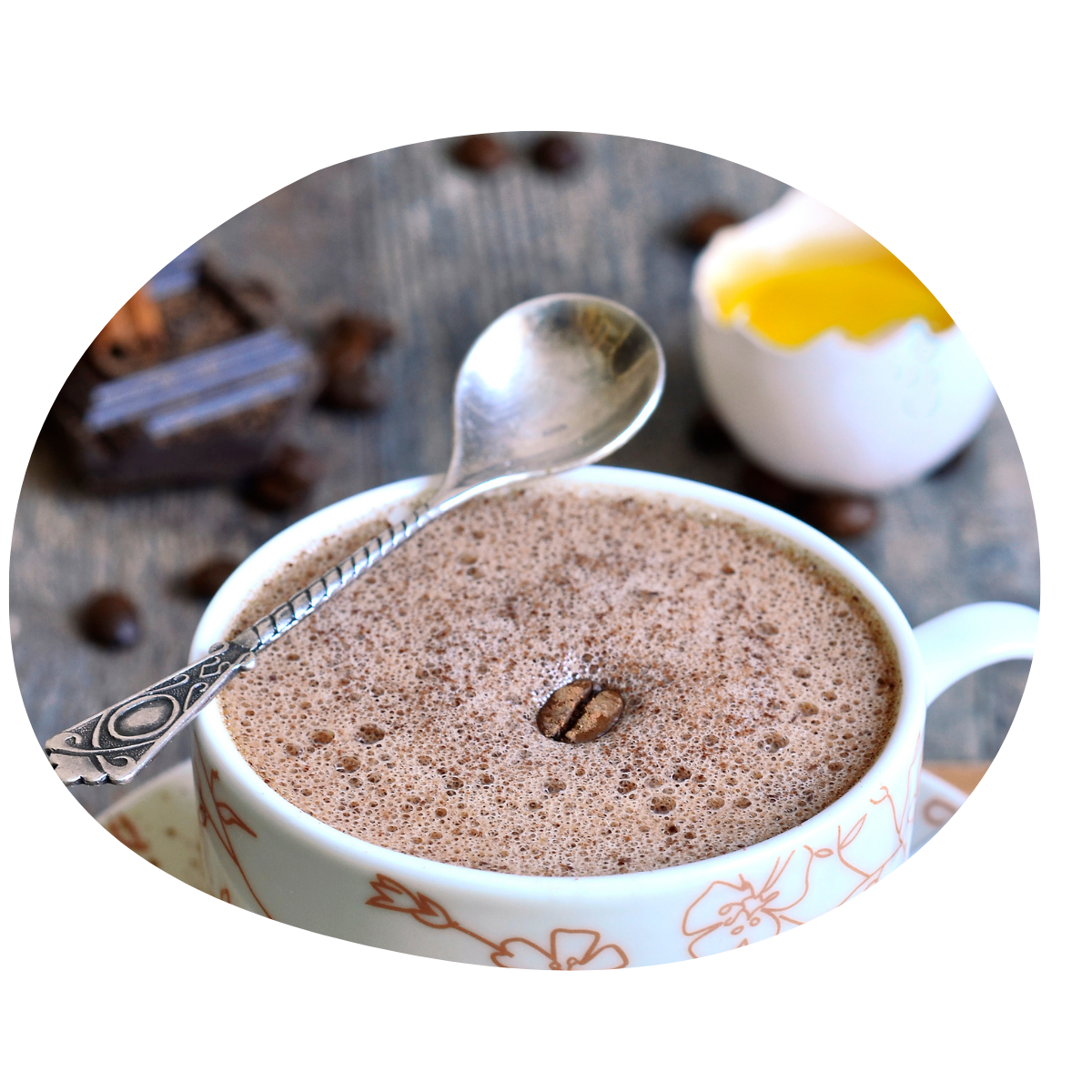 Eine Tasse Kaffee mit Löffel, dahinter eine geöffnete Eierschale mit Eidotter